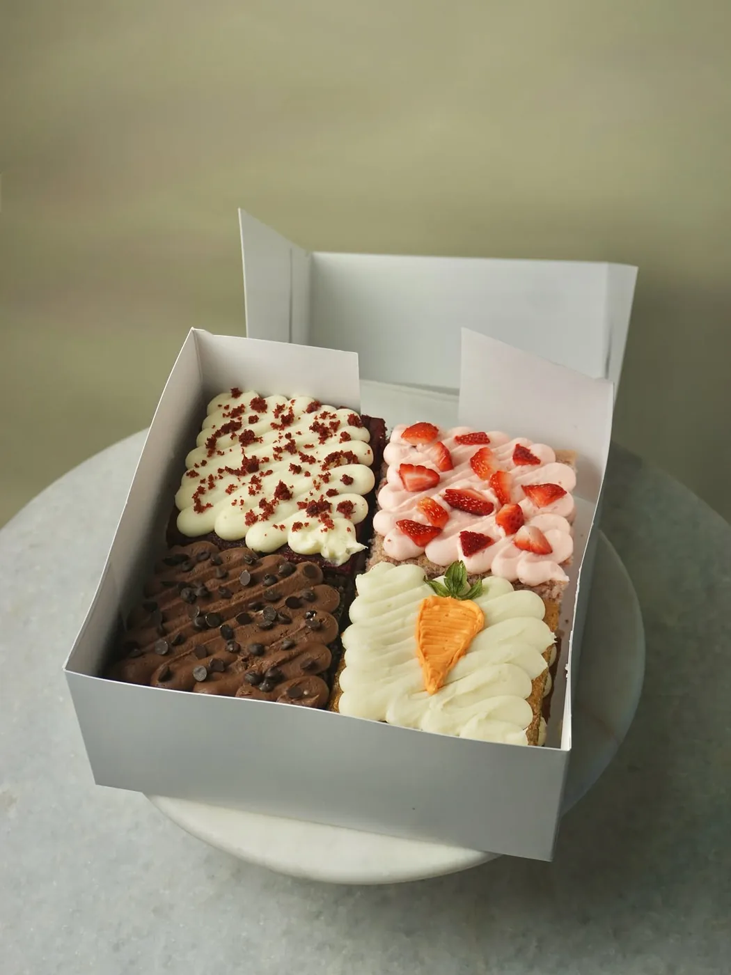 A white bakery box holding four decorated Morning Brew Co bars: one piped with chocolate buttercream and chocolate chips, one topped with sliced strawberries, one with red crumb, and one piped with a carrot