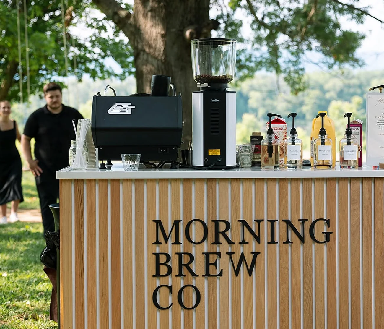 The Morning Brew Co coffee cart set up outdoors under a tree, espresso machine and grinder on the counter