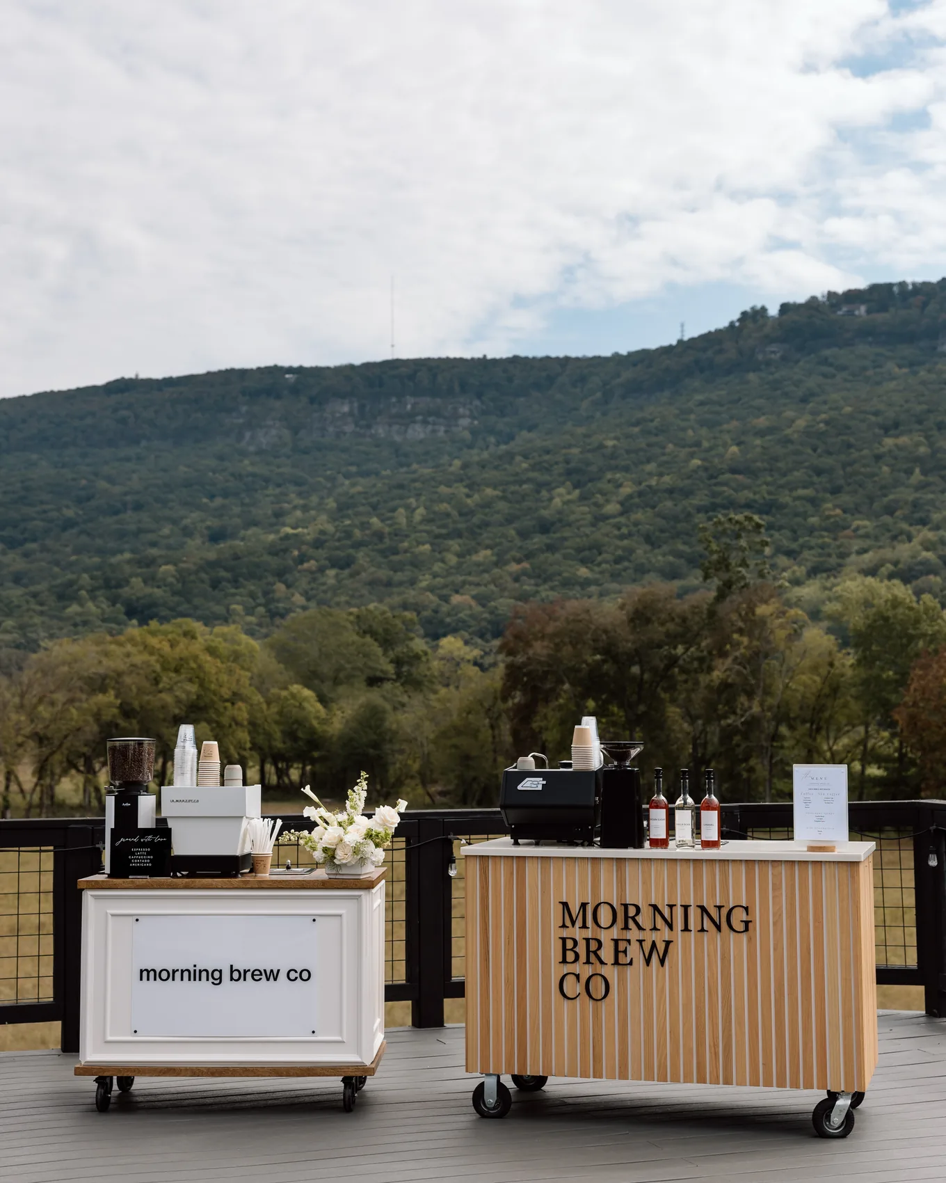 The Morning Brew Co coffee cart set up on a terrace, two branded units with the espresso machine, syrups and flowers, mountains behind