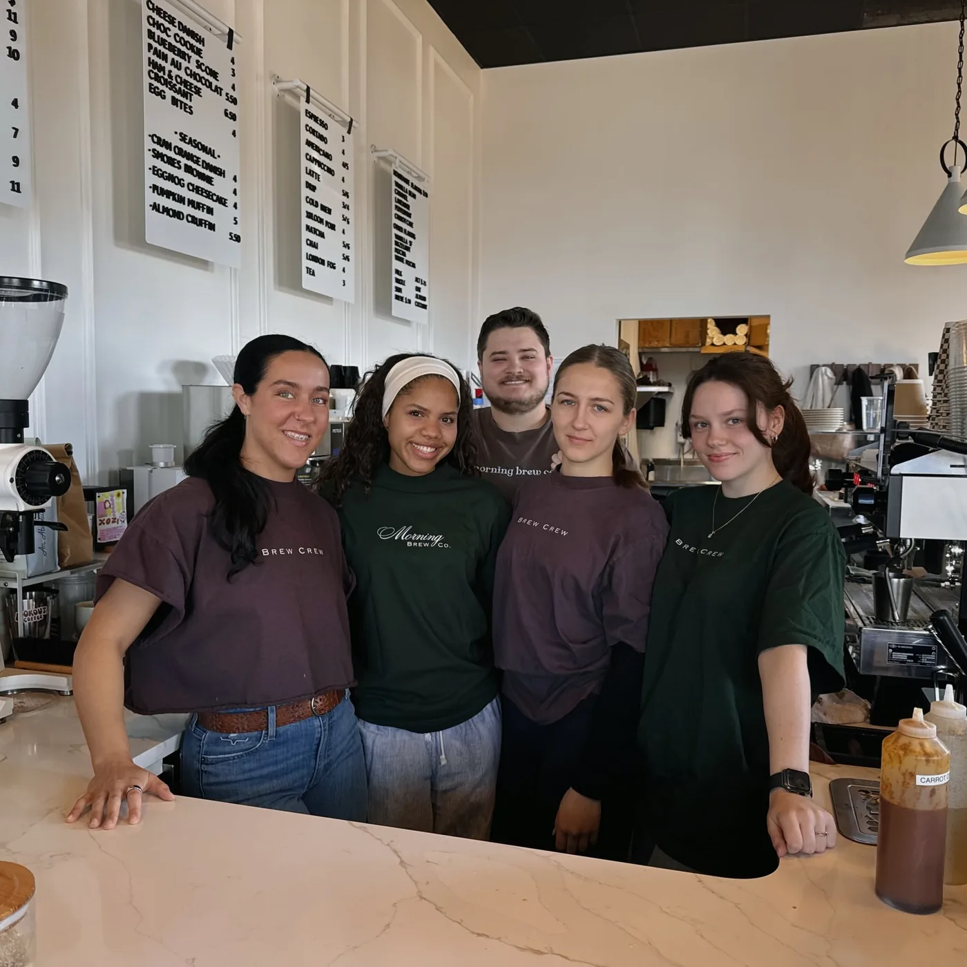 Five of the Morning Brew Co crew in brew crew shirts behind the marble counter, the printed menu boards and the espresso machine behind them