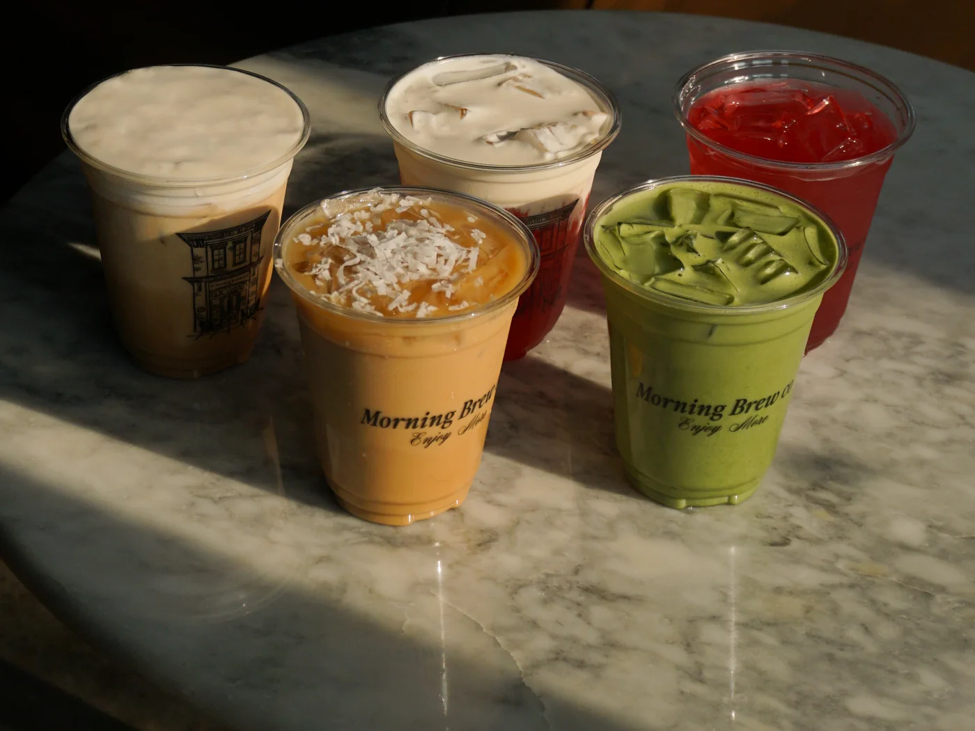 Five Morning Brew Co drinks on a marble table in afternoon light: two iced lattes, a coconut topped latte, a matcha and a red refresher
