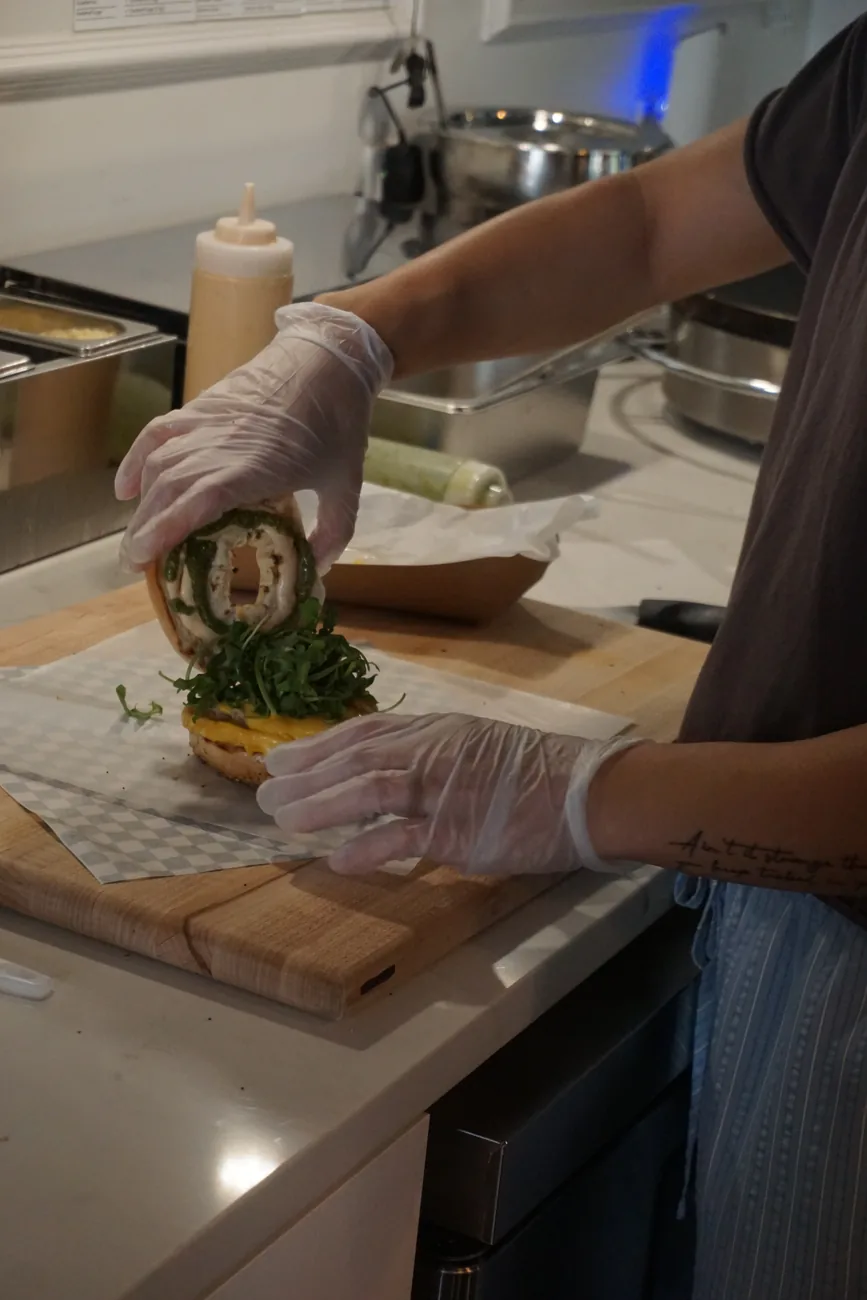 Gloved hands assembling a bagel sandwich on a board in the Morning Brew Co kitchen