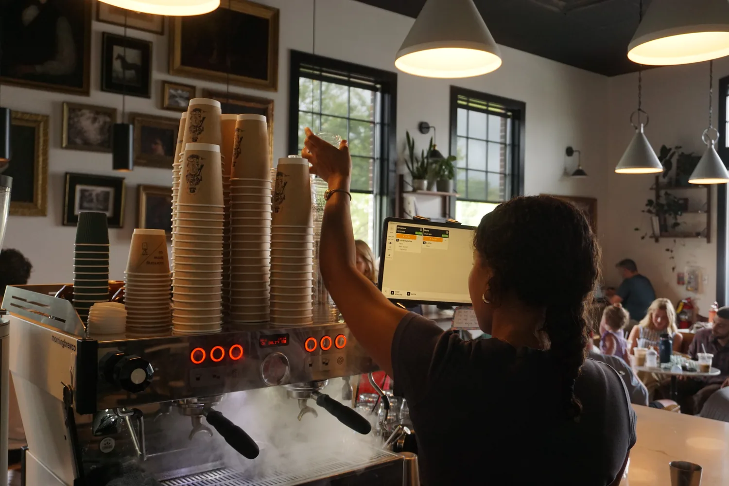 A barista reaching for cups above the espresso machine while the room fills behind