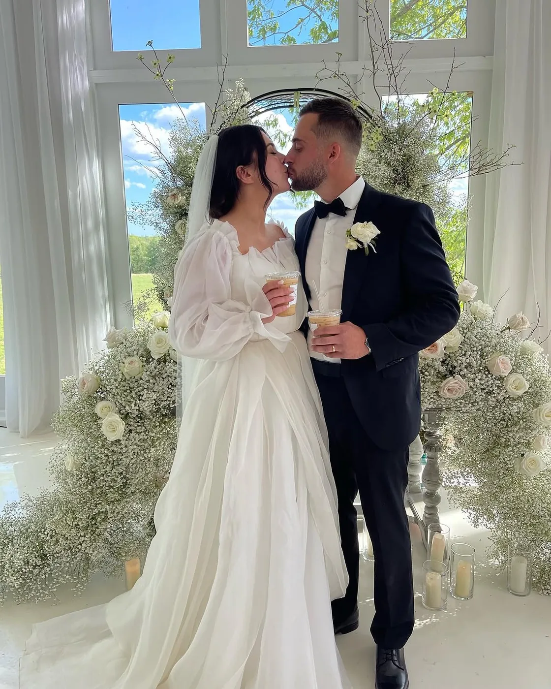 A bride and groom kissing in front of a white floral arch of roses and baby's breath, each holding a clear cup of iced coffee from the Morning Brew Co cart, tall windows and green trees behind